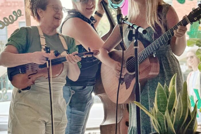 three piece band of women gathered around single mic