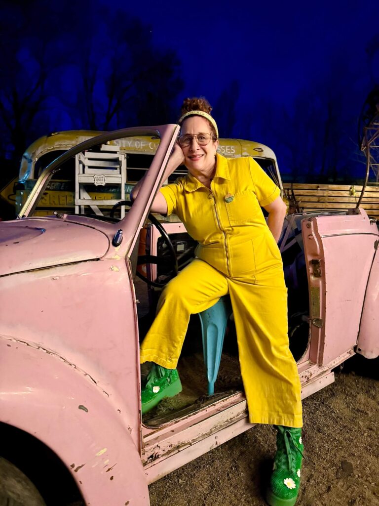 woman in yellow jumpsuit and green astroturf boots standing by pink VW bug convertible car