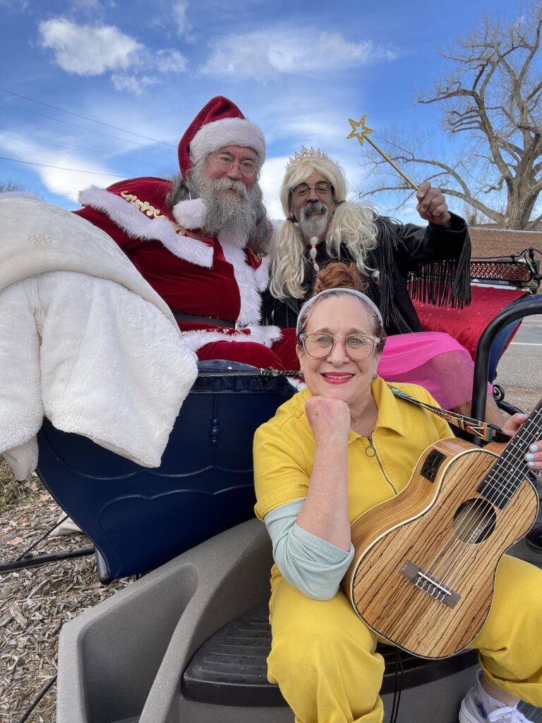woman in yellow jumpsuit with tiny guitar by sleigh with Santa and man with beard, leather jacket and pink tutu