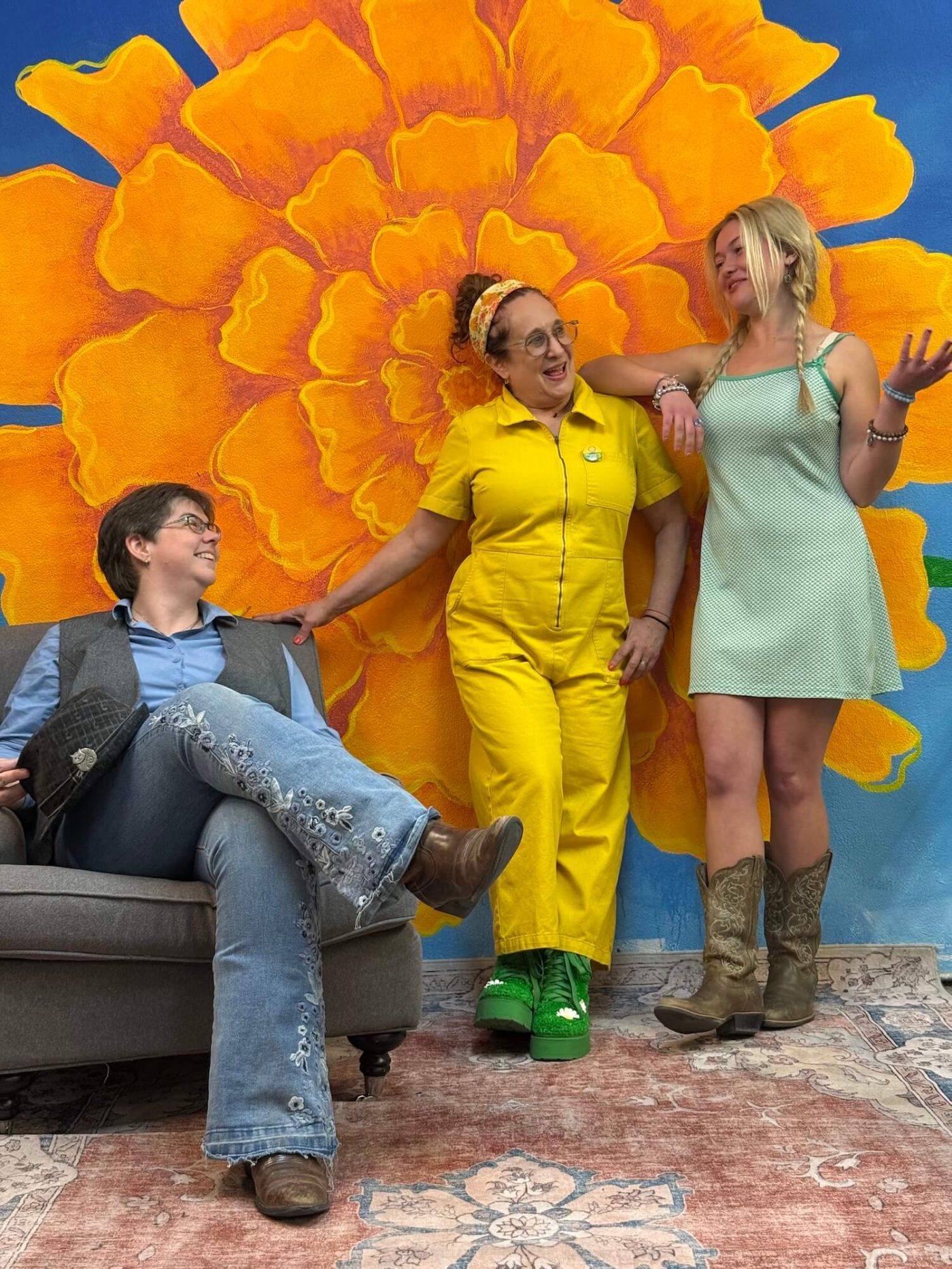 three women sitting and standing by large flower mural, smiling